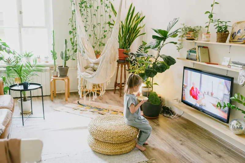 Child enjoying a snack in a cozy living room with lush plants and a hammock chair